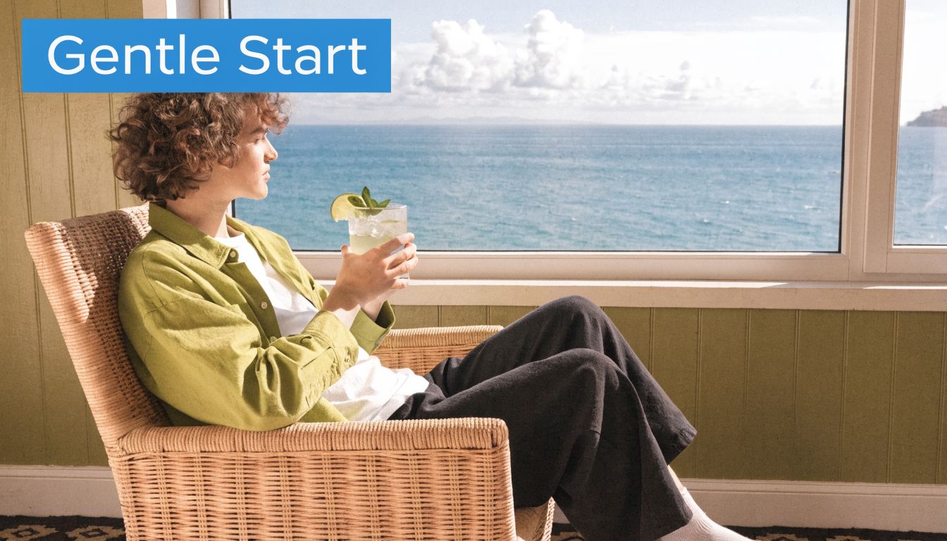 A young person with curly hair relaxes in a wicker chair by the sea, holding a refreshing drink.