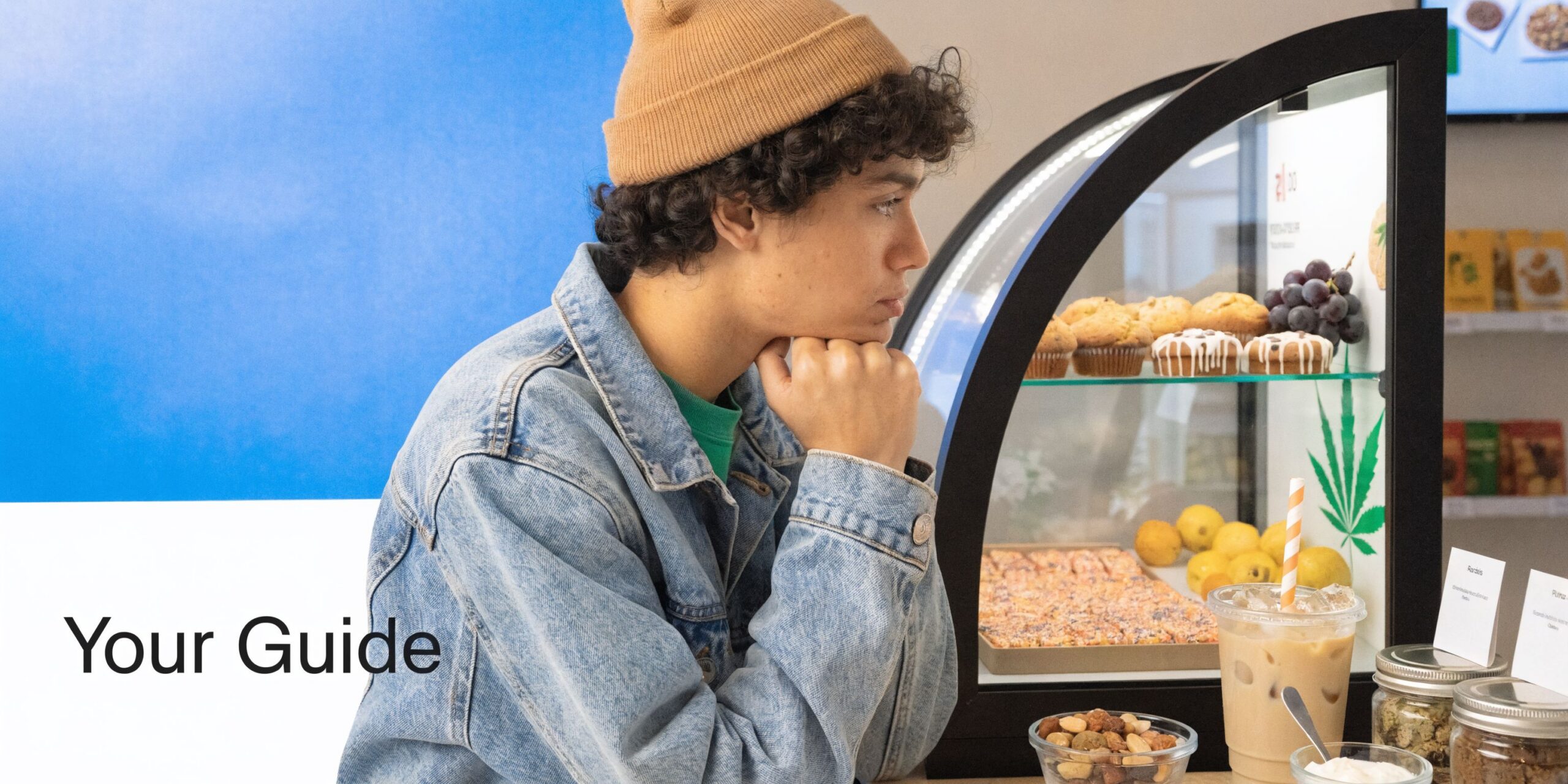 A young man wearing a beanie leans against a cafe counter, gazing at display case items.
